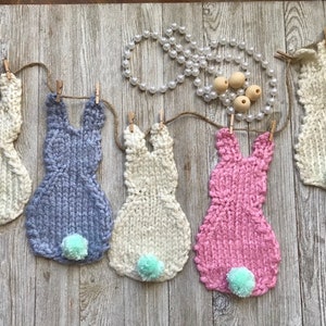 May include: A garland of five knitted bunny shapes, each with a fluffy tail. The bunnies are in white, light blue, pink, and two more white. The garland is strung with twine and wooden clothes pegs.