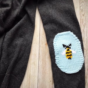 May include: Dark gray sweater with a light blue knitted elbow patch. The patch features a yellow and black bee with white wings. The sweater is made of a soft material and is suitable for casual wear.