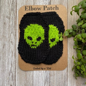 Op de afbeelding: Zwarte gebreide elleboogstukken met neon groene schedelontwerpen. De patches worden gepresenteerd op een bruine kaart met de tekst "Elbow Patch" en "Sealed by a Kris". Op een houten oppervlak.
