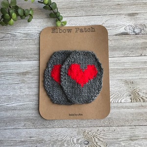 Puede incluir: Dos parches de codo de punto gris con un diseño de corazón rojo. Los parches están en una tarjeta marrón con el texto "Elbow Patch" y "Sewn by a Kira".