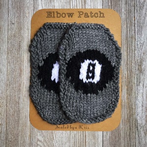 May include: Two gray knitted elbow patches with black and white circles. The patches are on a brown card with the text "Elbow Patch" and "Sealed by a Kris".
