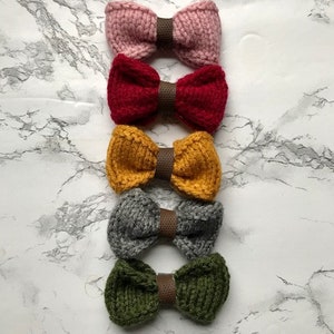 May include: Five knitted bow ties in different colours. The bow ties are arranged vertically, starting with pink, then red, yellow, grey, and green.