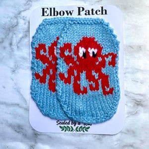 May include: Two blue knitted elbow patches with a red octopus design. The patches are on a white card with the text "ELBOW PATCH" at the top. The card also has the text "Sealed by a Kiss" and a leaf design.