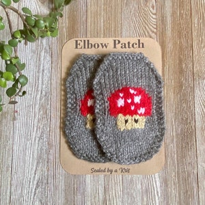 May include: Two gray knitted elbow patches with a red mushroom design. The patches are on a brown card with the text "Elbow Patch" and "Sealed by a Kris".