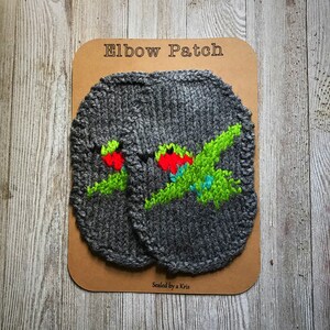 May include: Two gray knitted elbow patches with green, red, and blue embroidered designs. The patches are labeled "Elbow Patch" and "Sealed by a Kris".