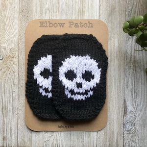 Könnte beinhalten: Zwei schwarze gestrickte Ellenbogen-Patches mit weißen Totenkopf-Designs. Die Patches befinden sich auf einer braunen Karte mit dem Text "Elbow Patch" und "Sealed by a Knitter".