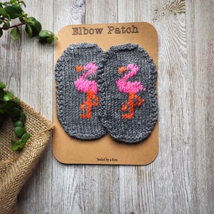 Peut inclure: Deux coudières en tricot gris avec des flamants roses et oranges brodés. Les patchs sont présentés sur une carte marron avec le texte "Elbow Patch". L'arrière-plan est une surface en bois avec de la verdure.