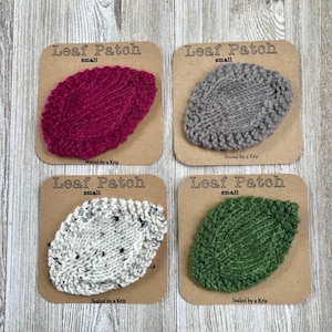 Może przedstawiać: Cztery małe dziane łatki w kształcie liści w różnych kolorach: bordowym, szarym, białym z czarnymi plamkami i zielonym. Każda łatka znajduje się na brązowej kartce z tekstem "Leaf Patch small" i "Sealed by a Kris".