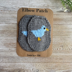 May include: Two gray knitted elbow patches with a blue bird design. The patches are packaged on a brown card with the text "Elbow Patch" and "Sealed by a Kris".