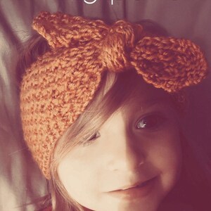 May include: A close-up of a child wearing a knitted headband with a bow. The headband is a warm orange color. The child is smiling and looking at the camera.
