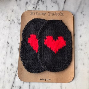 Puede incluir: Dos parches de codo de punto negro con diseños de corazón rojo. Los parches están empaquetados en una tarjeta marrón con el texto "Elbow Patch" y "Sealed by a Kris".