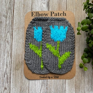May include: Two gray knitted elbow patches with a blue and green flower design. The patches are on a brown card with the text "Elbow Patch" and "Sealed by a Kris".