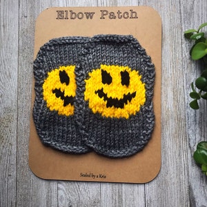 Puede incluir: Dos parches de codo de punto gris con caras sonrientes amarillas. Los parches están en una tarjeta marrón con el texto "Elbow Patch" y "Sealed by a Kris".
