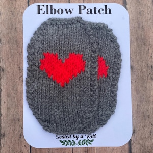 Puede incluir: Dos parches para codo tejidos en color gris oscuro con forma ovalada, cada uno con un diseño de corazón rojo brillante. Los parches están montados sobre una tarjeta blanca con el texto Elbow Patch arriba y Sealed by a Kris abajo.