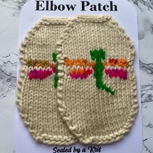 May include: Two beige, knitted elbow patches with a dragonfly design. The dragonfly has green, orange, and pink accents. The patches are on a white card with the text "Elbow Patch" and "Sealed by a Kris".