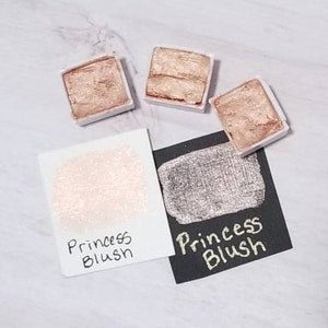 Pode incluir: Dois pequenos recipientes quadrados de tinta metálica com as palavras "Princess Blush" escritas nos rótulos. Um recipiente é de cor rosa claro e o outro é de cor prateada.