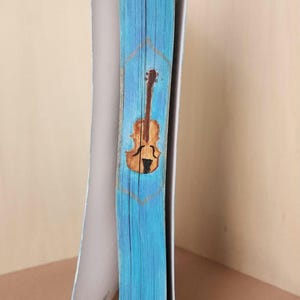 Op de afbeelding: Een boek met een lichtblauwe rug met een handgeschilderd vioolontwerp. De viool is bruin met een zwart klankgat, geplaatst in een goudkleurig geometrisch frame. De pagina's zijn wit en het boek is open.