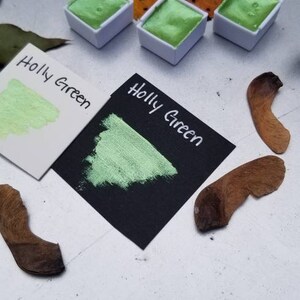 May include: Three small, white pans of green watercolor paint. The pans are labeled "Holly Green". A swatch of the paint is shown on a white card and a black card.