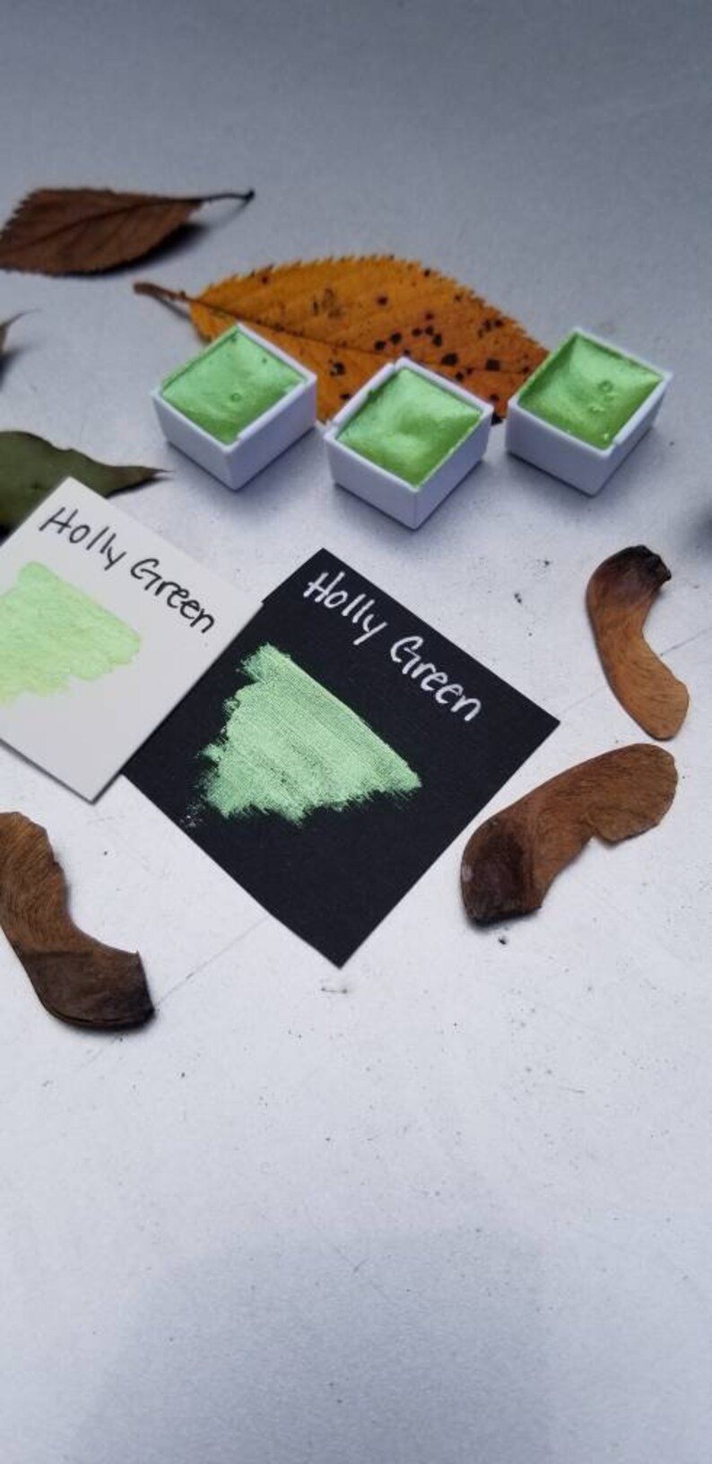 May include: Three small, white pans of green watercolor paint. The pans are labeled "Holly Green". A swatch of the paint is shown on a white card and a black card.