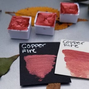 May include: Close-up of watercolor paints in white containers. The paint is a metallic copper color. Two cards display the words "Copper Fire" and swatches of the paint. The background includes a leaf and a neutral surface.
