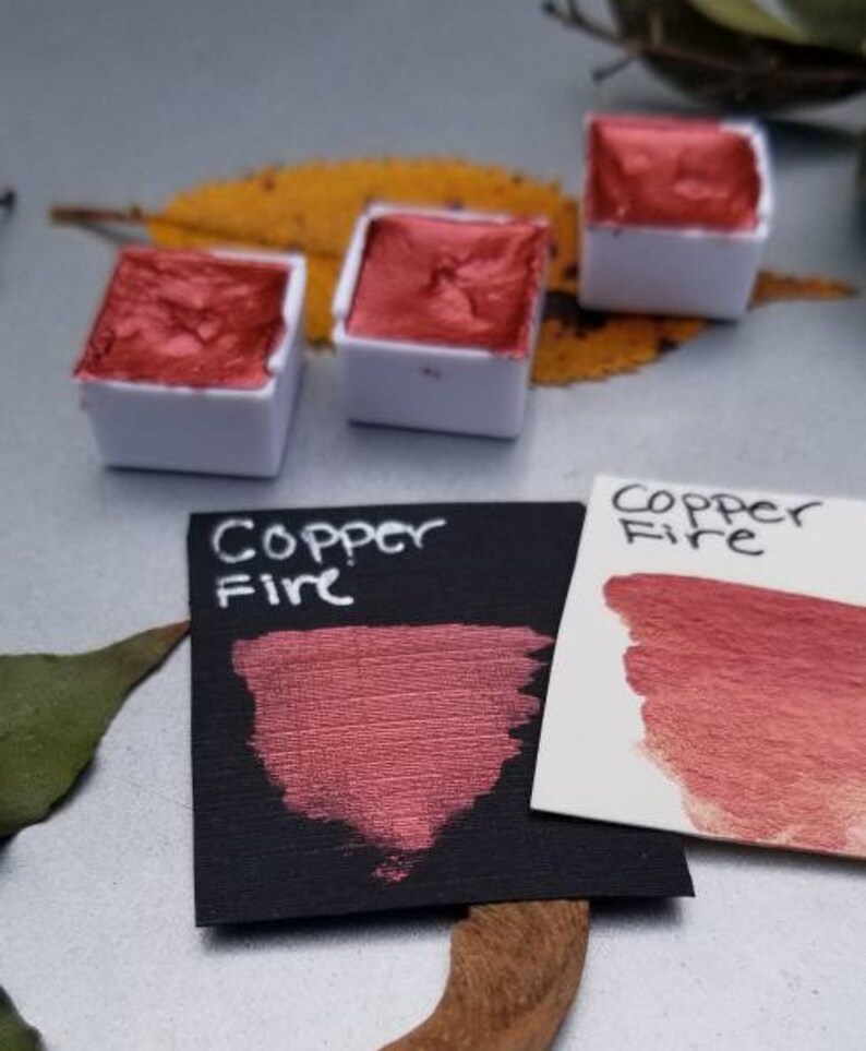 May include: Close-up of watercolor paints in white containers. The paint is a metallic copper color. Two cards display the words "Copper Fire" and swatches of the paint. The background includes a leaf and a neutral surface.