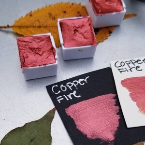 May include: Three small, white watercolor paint pans filled with metallic copper-colored paint. A swatch of the paint is on a black card with the words "Copper Fire" written in white. A leaf is in the background.