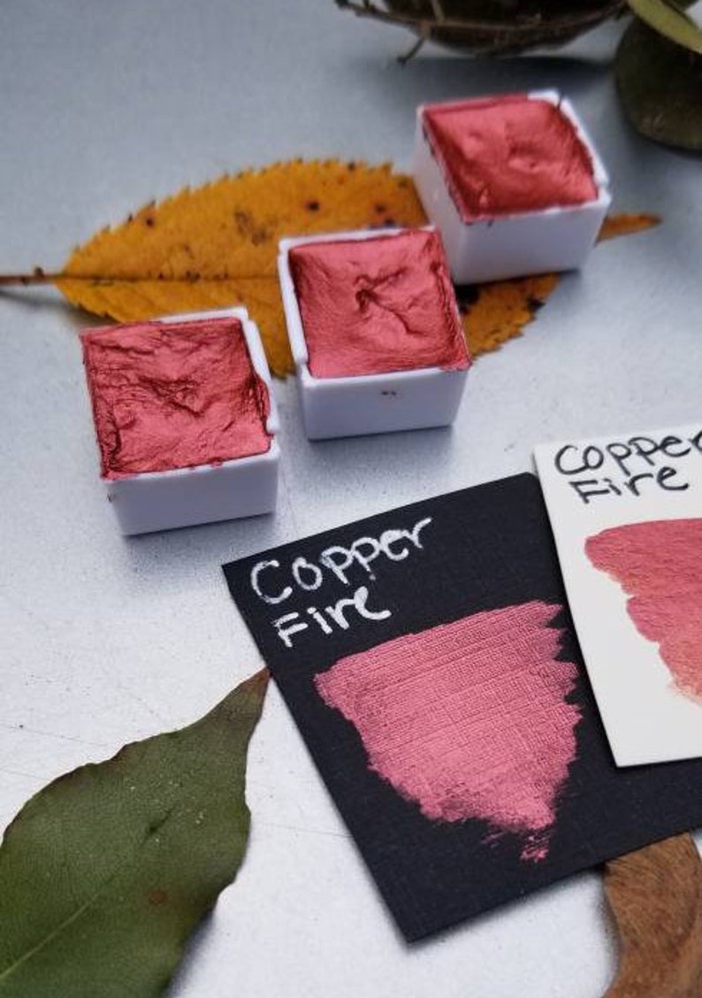 May include: Three small, white watercolor paint pans filled with metallic copper-colored paint. A swatch of the paint is on a black card with the words "Copper Fire" written in white. A leaf is in the background.