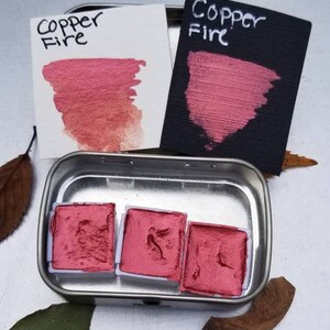 May include: A metal tin containing three square watercolor paint pans and two color swatches. The swatches and paint are a metallic copper-red color. The text "Copper Fire" is written on the swatches in black ink. The paint is a shimmery, metallic finish.