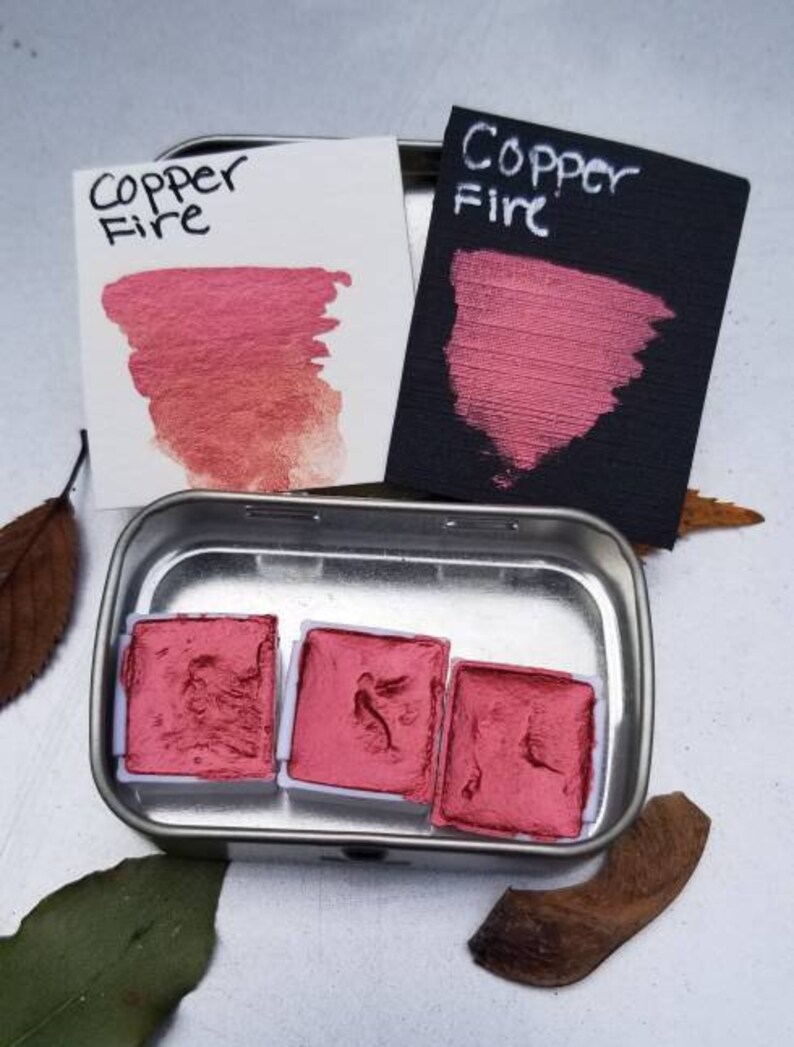 May include: A metal tin containing three square watercolor paint pans and two color swatches. The swatches and paint are a metallic copper-red color. The text "Copper Fire" is written on the swatches in black ink. The paint is a shimmery, metallic finish.