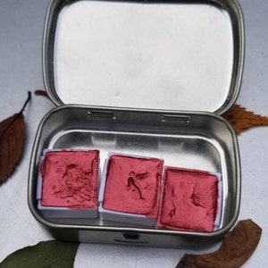 May include: Open, silver-colored metal tin containing three square, red watercolor paint pans. The paint is a deep, rich red. The tin is open, revealing the paint inside. The background is a neutral gray.