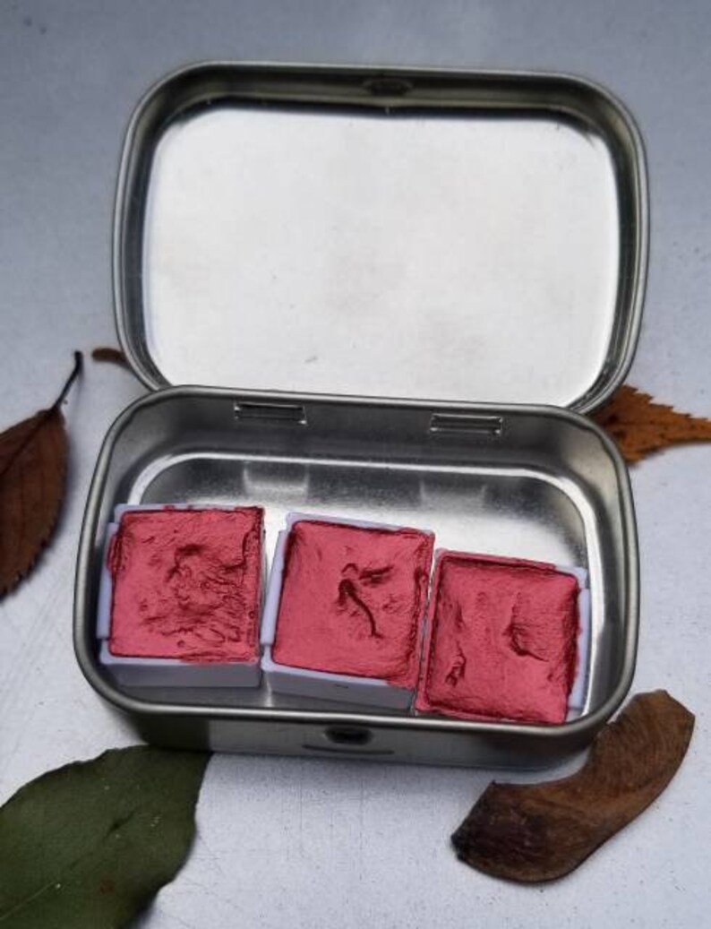 May include: Open, silver-colored metal tin containing three square, red watercolor paint pans. The paint is a deep, rich red. The tin is open, revealing the paint inside. The background is a neutral gray.