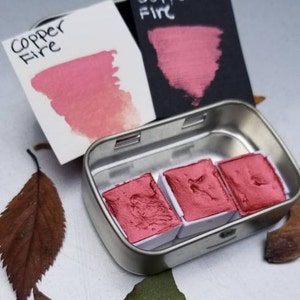 May include: A small metal tin containing three rectangular watercolor paint pans in a vibrant copper-red hue. Two color swatches, one on white and one on black, display the name "Copper Fire". The image includes fallen leaves.