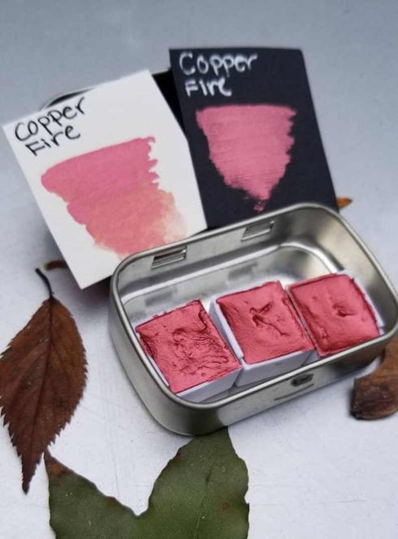 May include: A small metal tin containing three rectangular watercolor paint pans in a vibrant copper-red hue. Two color swatches, one on white and one on black, display the name "Copper Fire". The image includes fallen leaves.