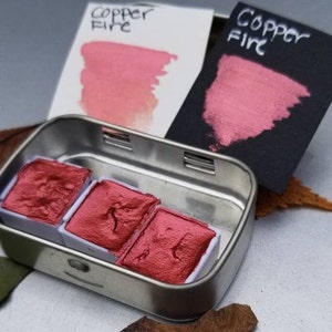 May include: A metal tin containing three red watercolor paint pans. Two cards with the text "Copper Fire" are behind the tin, displaying swatches of the paint. One card is white, the other is black. The paint has a metallic sheen.