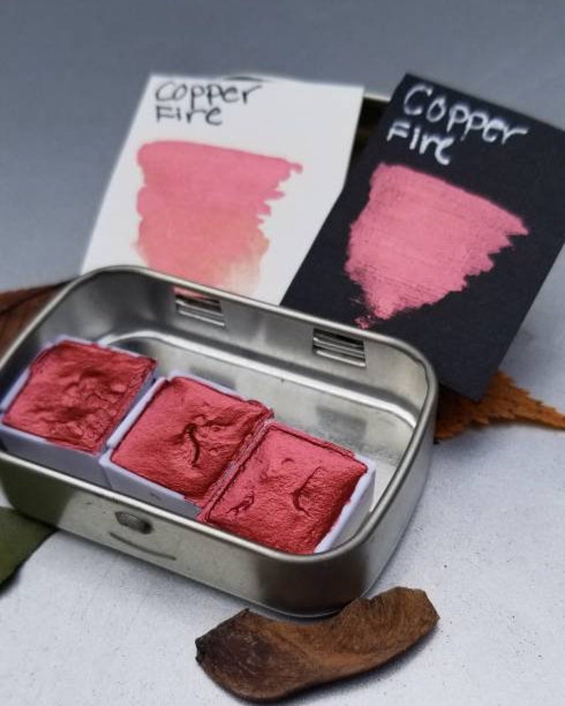 May include: A metal tin containing three red watercolor paint pans. Two cards with the text "Copper Fire" are behind the tin, displaying swatches of the paint. One card is white, the other is black. The paint has a metallic sheen.