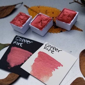 May include: Three square watercolor pans filled with metallic copper-red paint sit next to two color swatches. The swatches are labeled "Copper Fire" and show the paint's color on black and white paper. The image has a natural, artistic theme.