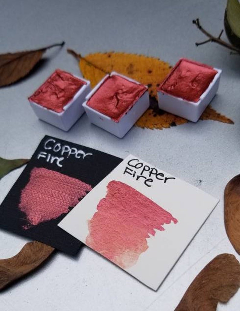 May include: Three square watercolor pans filled with metallic copper-red paint sit next to two color swatches. The swatches are labeled "Copper Fire" and show the paint's color on black and white paper. The image has a natural, artistic theme.