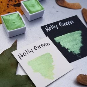 May include: Three watercolor paint pans with a green pigment labeled "Holly Green". The paint is a metallic green with a shimmery finish. The pans are on a white surface with a leaf and other natural elements.