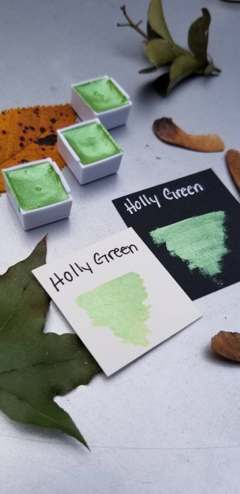 May include: Three watercolor paint pans with a green pigment labeled "Holly Green". The paint is a metallic green with a shimmery finish. The pans are on a white surface with a leaf and other natural elements.