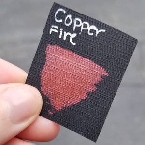May include: A small, black rectangular card with the words "Copper Fire" in white script. A swatch of metallic red paint is brushed onto the card, showcasing the color. The card is held by a person's fingers.