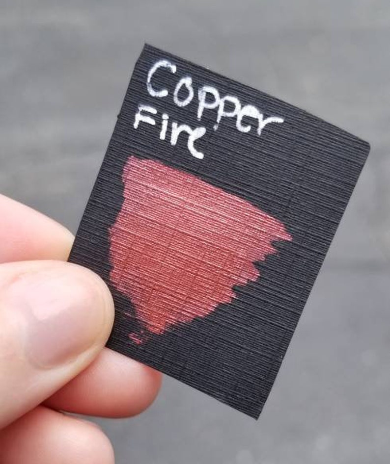 May include: A small, black rectangular card with the words "Copper Fire" in white script. A swatch of metallic red paint is brushed onto the card, showcasing the color. The card is held by a person's fingers.