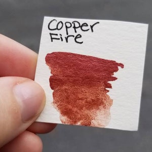 May include: A small white card with the words "Copper Fire" written in black ink. A swatch of metallic copper-red paint is brushed onto the card, showing color variations from dark to light.