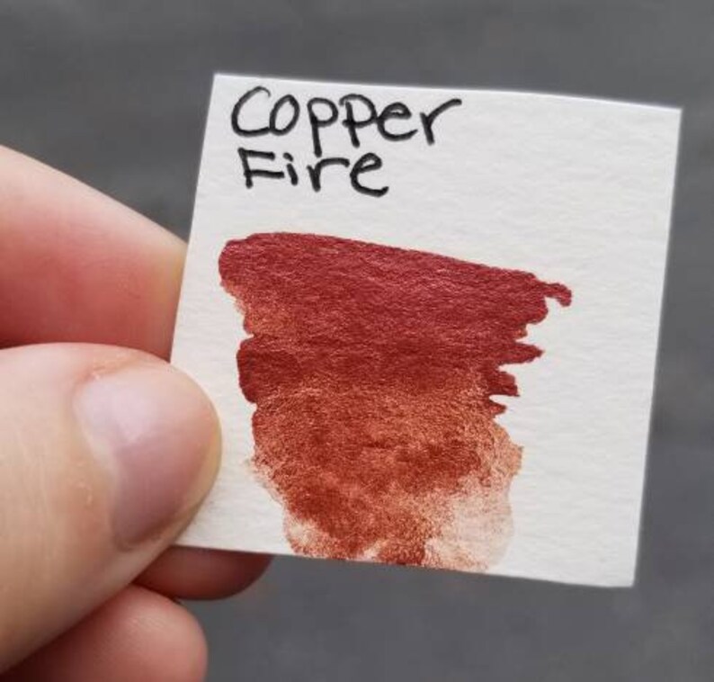 May include: A small white card with the words "Copper Fire" written in black ink. A swatch of metallic copper-red paint is brushed onto the card, showing color variations from dark to light.
