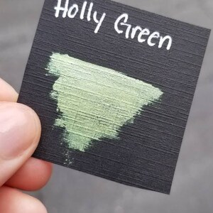 May include: A black card with the words "Holly Green" written in white. A swatch of light green paint is on the card.