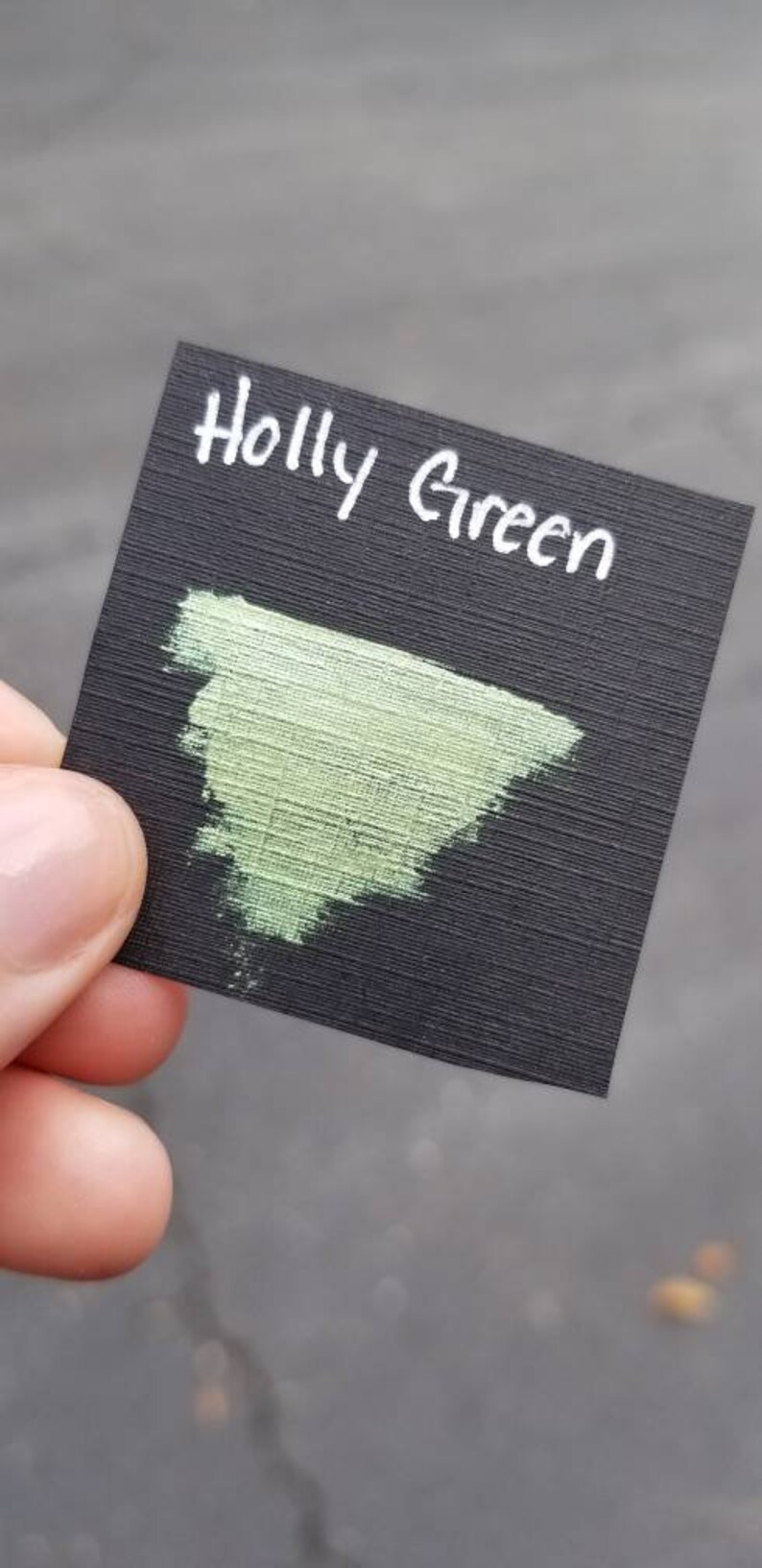 May include: A black card with the words "Holly Green" written in white. A swatch of light green paint is on the card.