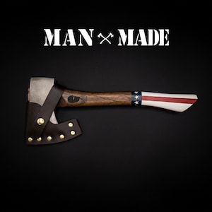 May include: A hatchet with a metal head, a dark brown wooden handle, and a leather sheath secured with brass rivets. The handle features a blue band with white stars and red and white stripes, and the text MAN MADE is printed above.