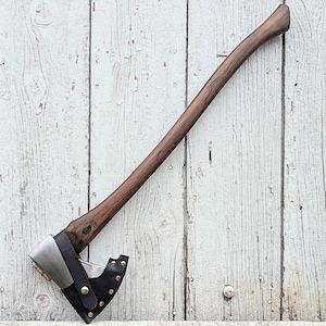 May include: Axe with a long, dark wooden handle and a silver and black head. The axe head is protected by a black leather sheath secured with brass-colored rivets. The axe is set against a weathered, white wooden background.