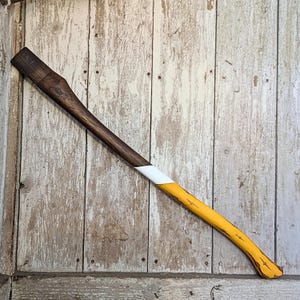 May include: A wooden axe handle painted with yellow and white paint. The handle is brown and has a slight curve at the end.