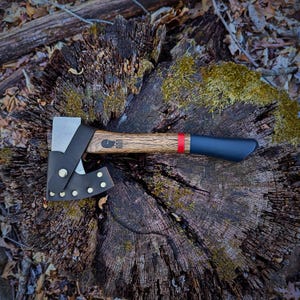 May include: A small axe with a black handle and a red stripe. The axe has a leather sheath with brass rivets. The axe is resting on a tree stump.