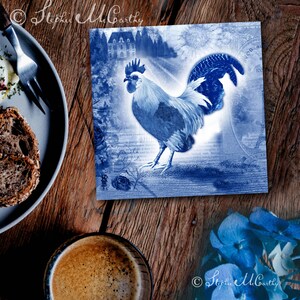 May include: A blue and white rooster illustration on a tile with a vintage, distressed look. The rooster is standing on a path with a building in the background. The tile has a blue and white floral design around the edges.